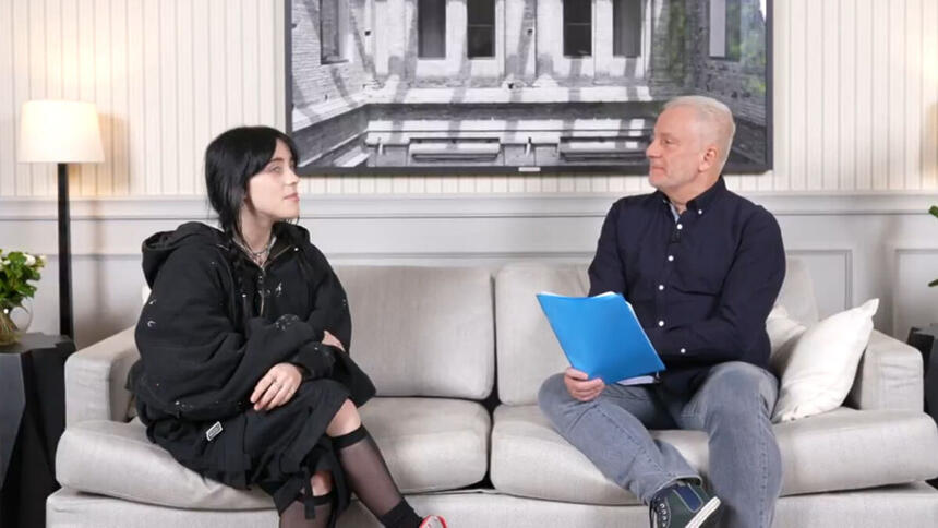 Billie Eilish em frente ao repórter Alberto Pereira Jr. sentados em um sofá, em trecho da entrevista para o Fantástico