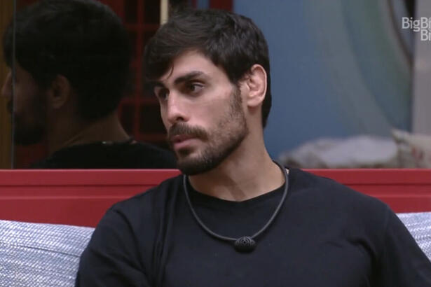 Cara de Sapato sentado na sala do BBB 23, vestindo uma camiseta preta e olhando para o lado