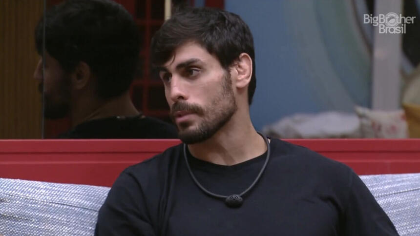 Cara de Sapato sentado na sala do BBB 23, vestindo uma camiseta preta e olhando para o lado