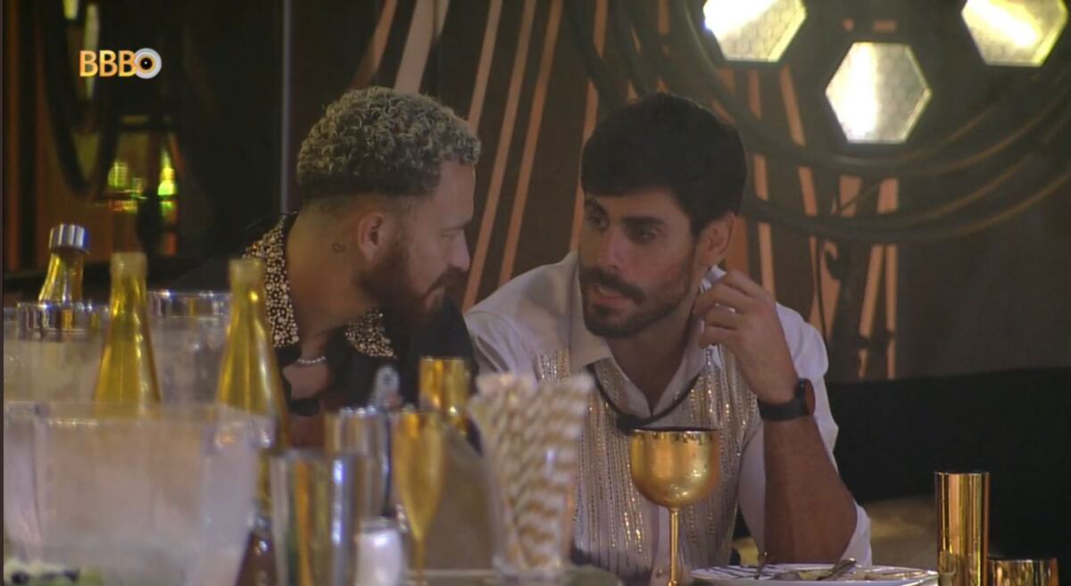 Fred conversa com Cara de Sapato na festa. Os dois estão numa mesa que se encontra com copos e bebidas.