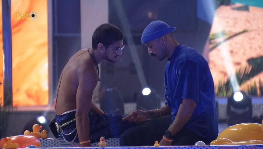 Gabriel sentado de frente para Ricardo na área externa do BBB 23. O ator está sem camisa e com uma calça. O biomédico está com uma boina azul na cabeça e uma camisa azul.