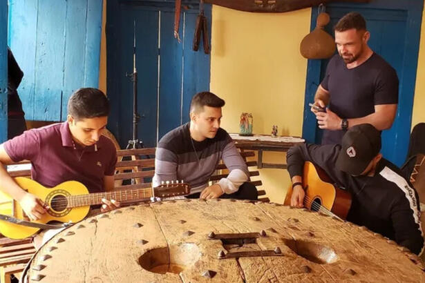 Fábio Castro em trecho do Globo Repórter, junto de cantores tocando violão em frente uma mesa