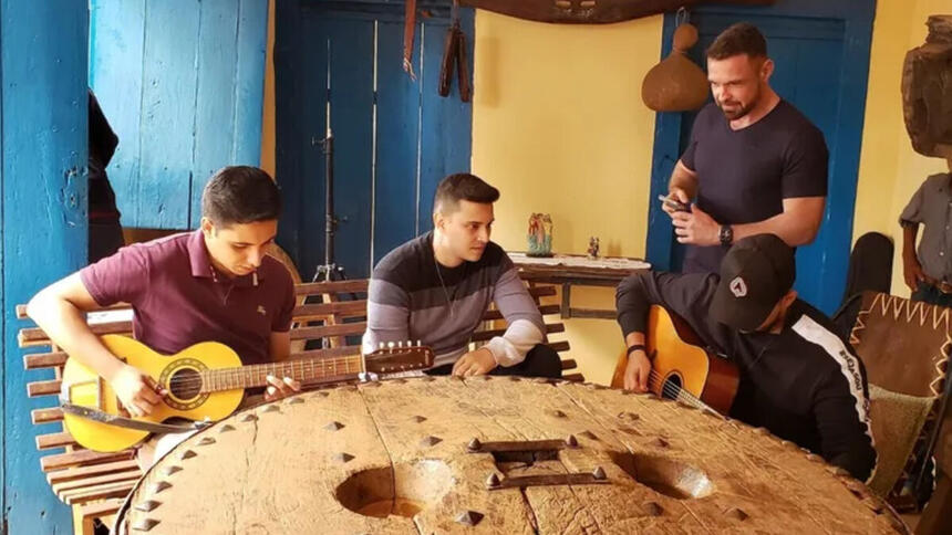 Fábio Castro em trecho do Globo Repórter, junto de cantores tocando violão em frente uma mesa