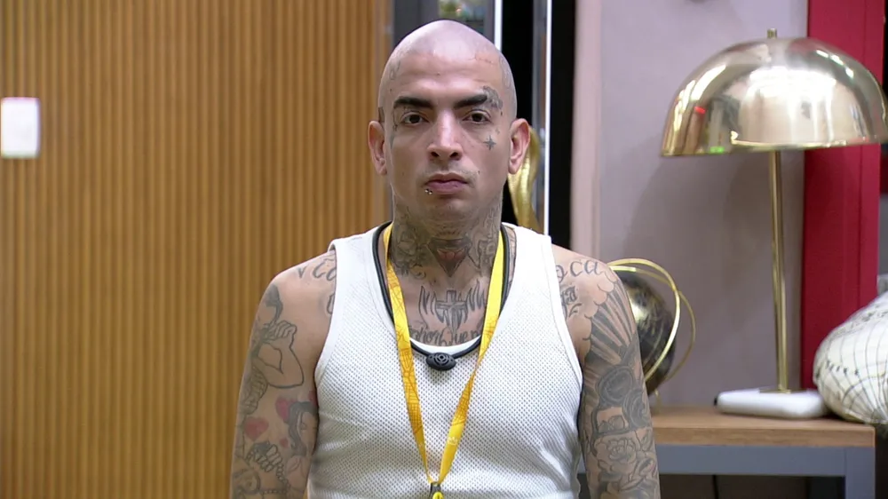 MC Guimê careca, de camisa regata branca, se olhando no espelho