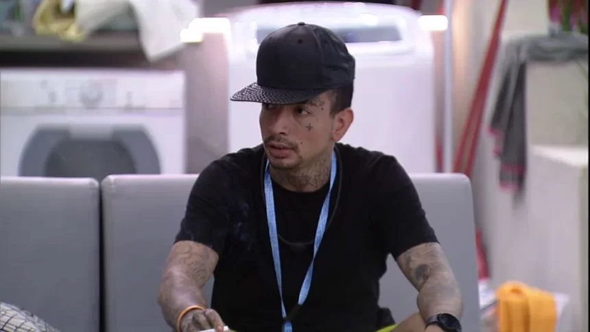 MC Guimê de chapéu e camisa preta, colar azul, sentado na varanda do BBB 23