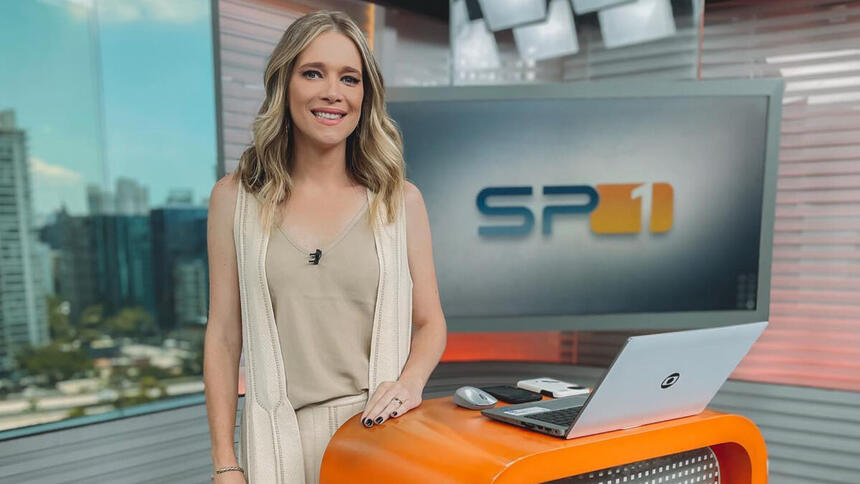 Natália Ariede em frente à bancada, no estúdio do SPTV, com o televisão ao fundo