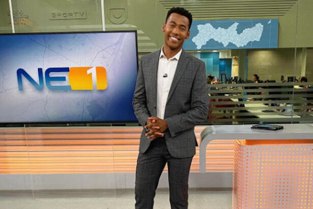 Pedro Lins posando no estúdio do NE1, com um televisor e a redação ao fundo