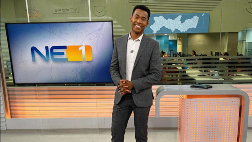 Pedro Lins posando no estúdio do NE1, com um televisor e a redação ao fundo