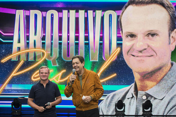 Rubens Barrichello ao lado de Faustão no centro do cenário da Band. Ao fundo o telão com o seu rosto e a arte do Arquivo Pessoal