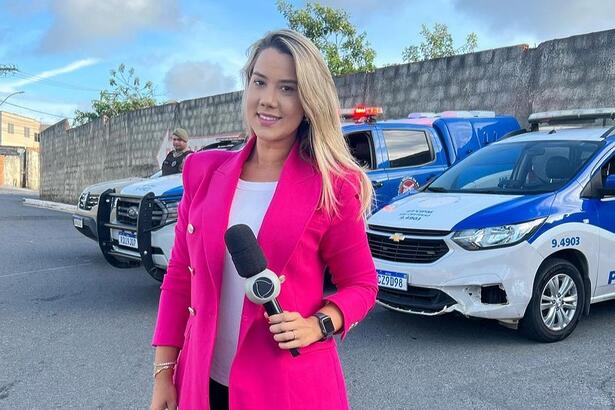 Repórter Daniela Mazzei segurando o microfone da Record TV Itapoan. Atrás estão carros da Polícia