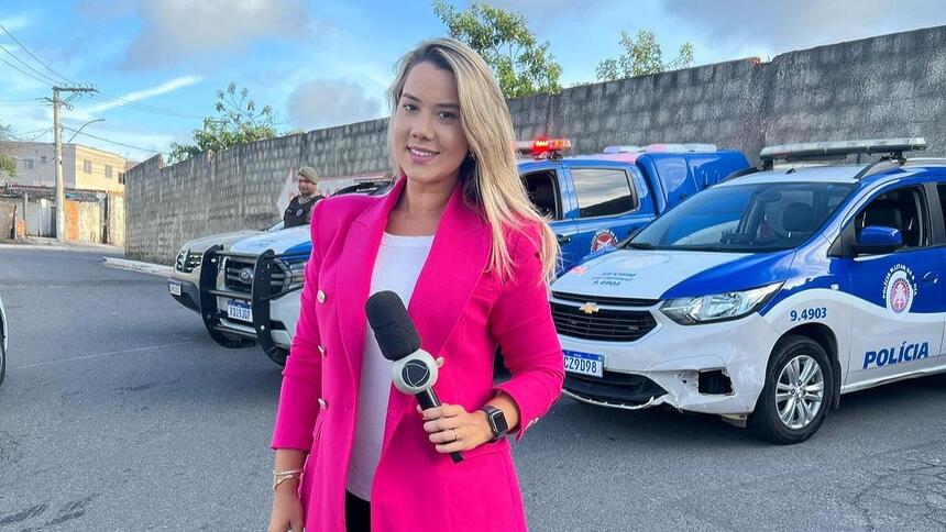 Repórter Daniela Mazzei segurando o microfone da Record TV Itapoan. Atrás estão carros da Polícia