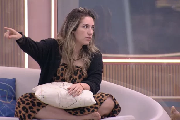 Amanda de vestido e sobretudo preto apontando pro lado direito, sentada no sofá da varanda do BBB 23