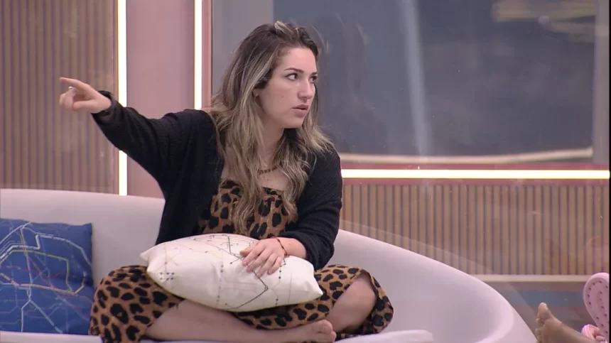 Amanda de vestido e sobretudo preto apontando pro lado direito, sentada no sofá da varanda do BBB 23