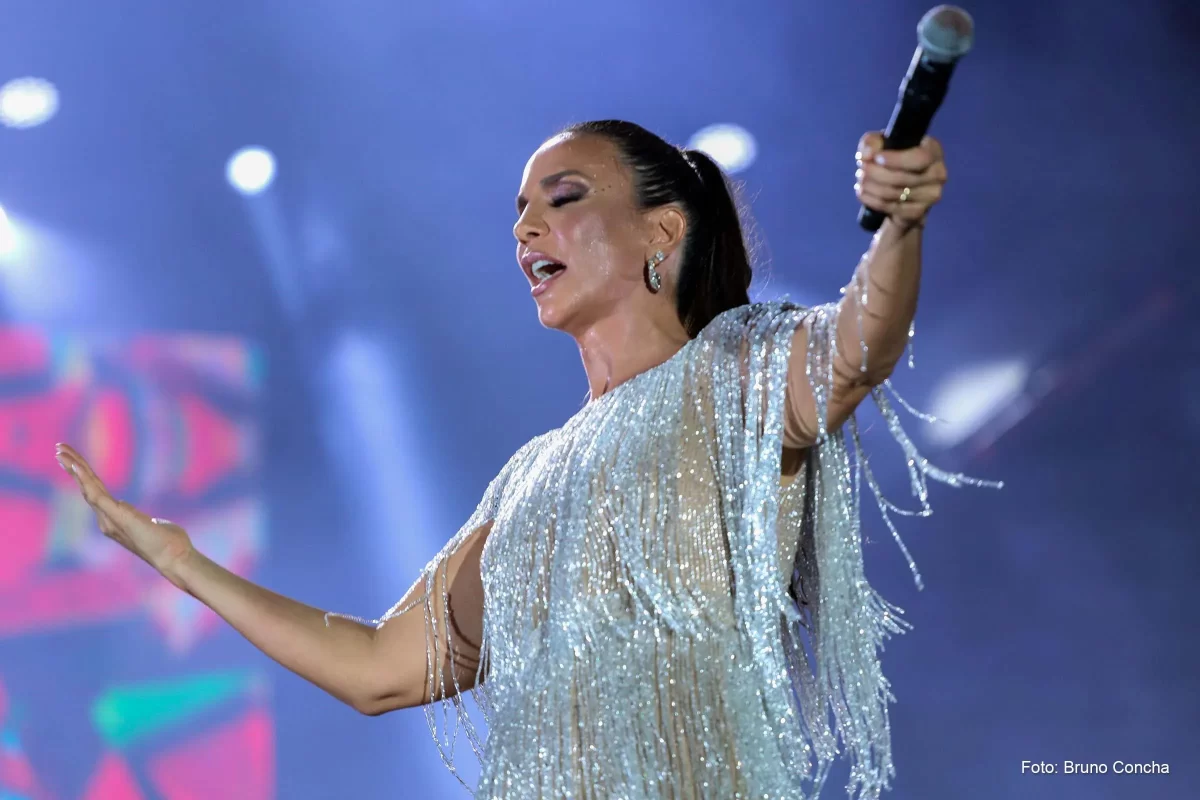 Ivete Sangalo com vestido brilhante, branco, com braços abertos segurando o microfone durante show na Bahia