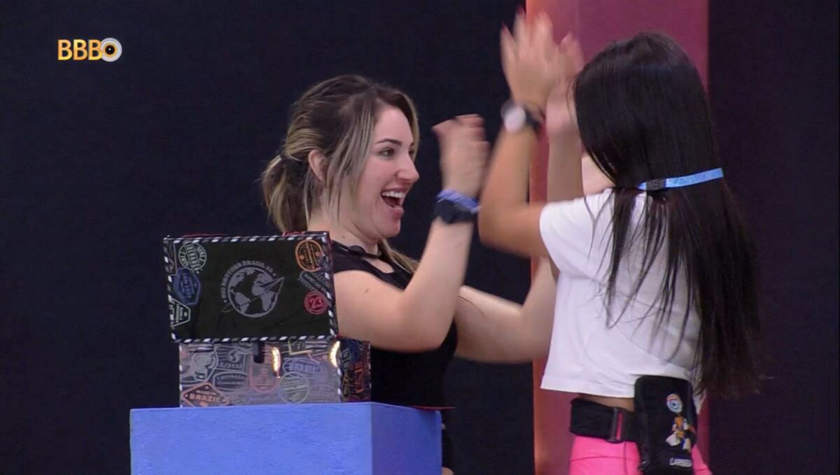 Amanda de frente para Larissa colocando um colar no seu pescoço.