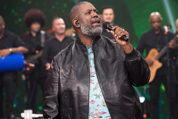 Péricles de jaqueta de couro e camisa azul, segudando o microfone durante gravação do Faustão na Band