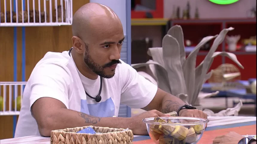 Ricardo de camisa branca, com semblante sério, com as mãos na mesa da cozinha do VIP do BBB 23