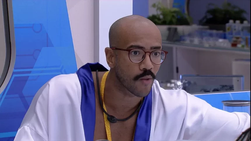 Ricardo, do BBB 23, com roupão branco de Líder, usando óculos de grau com o colar amarelo, sentado no Quarto do Líder