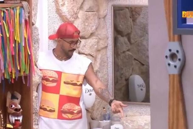 Ricardo em pé na porta do banheiro. O brother está com um chapéu na cabeça e vestido com uma camiseta regata estampada com desenhos de hambúrguer