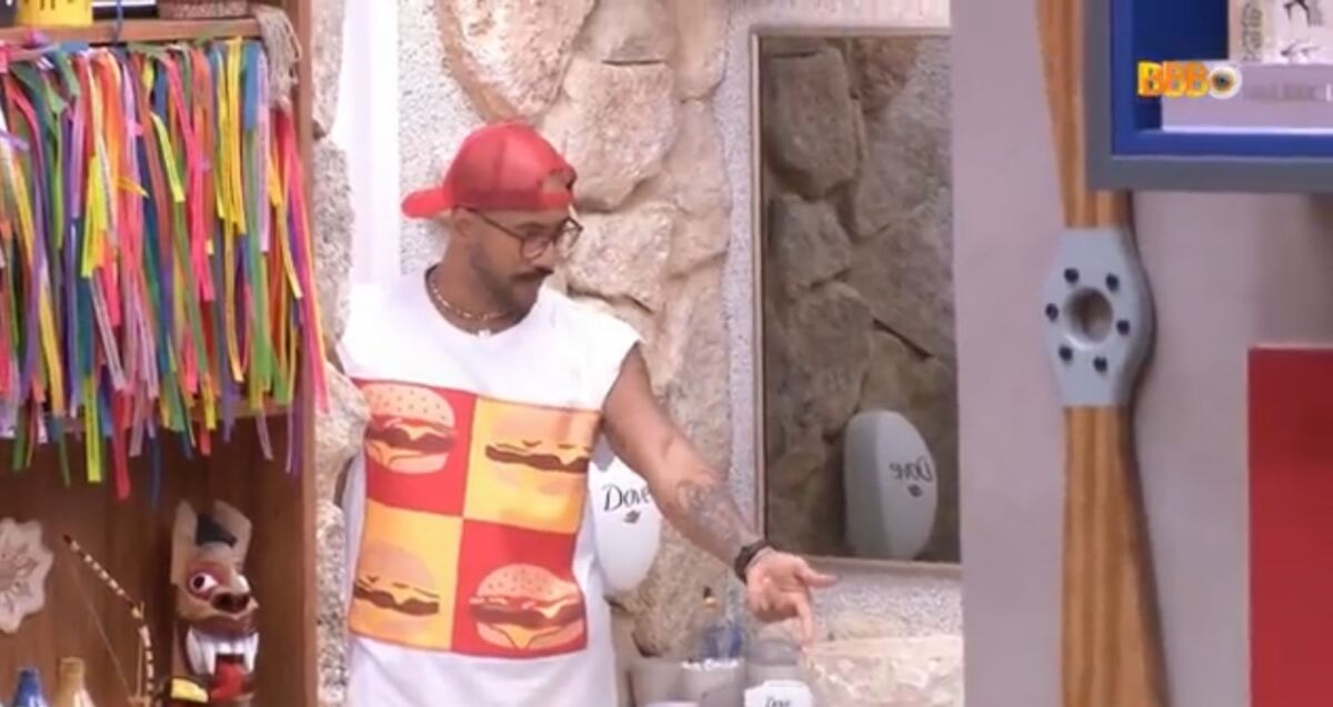 Ricardo em pé na porta do banheiro. O brother está com um chapéu na cabeça e vestido com uma camiseta regata estampada com desenhos de hambúrguer