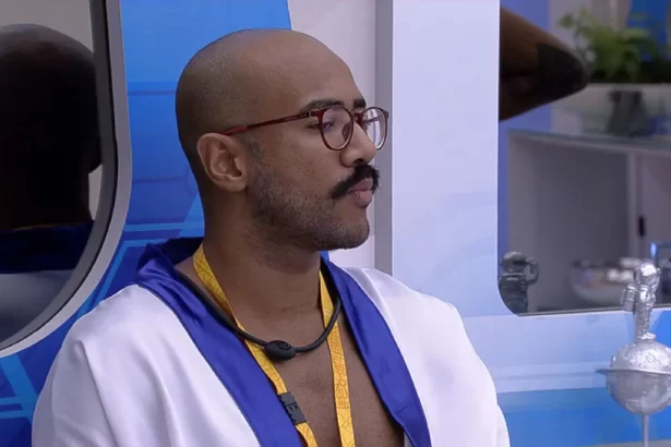 Ricardo, do BBB 23, com roupão branco de Líder, usando óculos de grau com o colar amarelo, sentado no Quarto do Líder