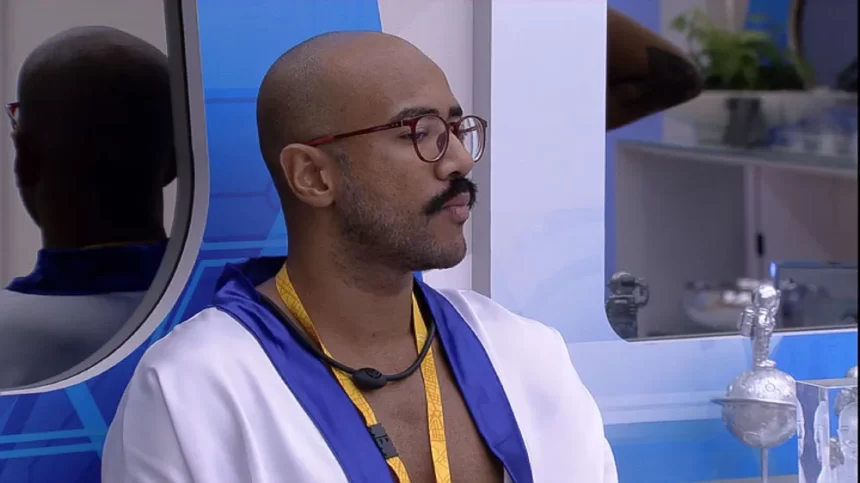 Ricardo, do BBB 23, com roupão branco de Líder, usando óculos de grau com o colar amarelo, sentado no Quarto do Líder