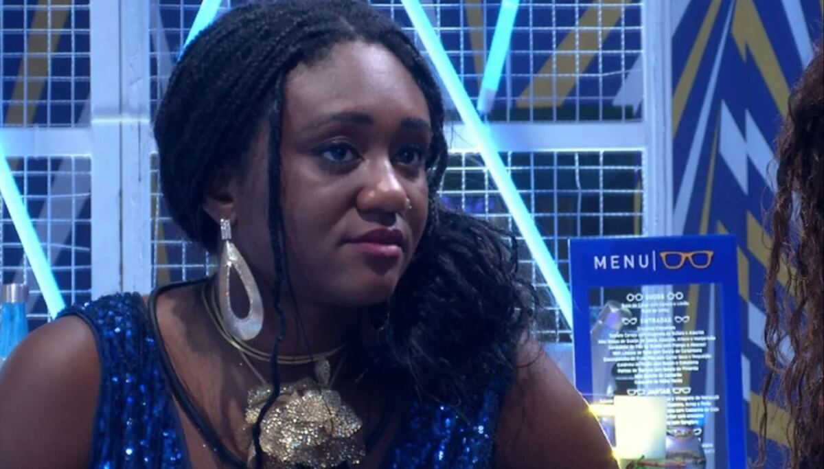 Sarah com semblante sério vestida com uma roupa azul brilhante com colar e brincos