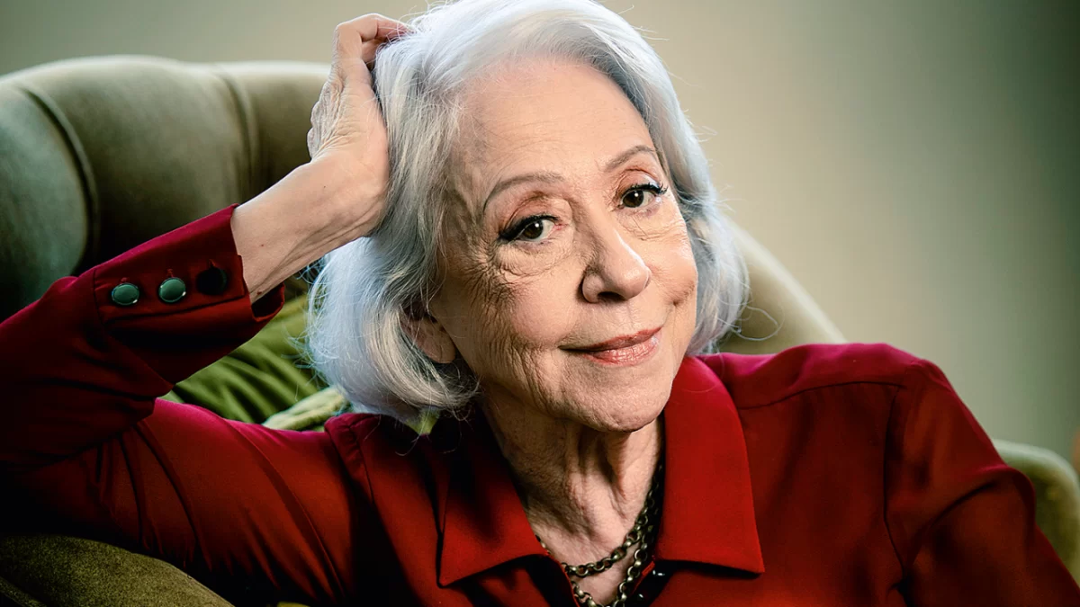 Fernanda Montenegro com a mão direita na cabeça, cabelo branco, solto, com camisa vermelha