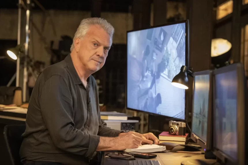 Pedro Bial de lado, sentado em frente a um monitor de tv, num birô com ferramentas de trabalho