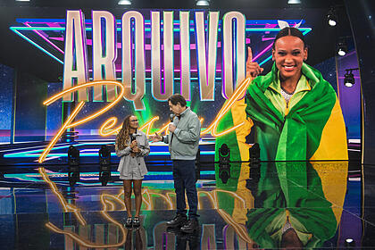 Rebeca Andrade olha para Faustão no centro do palco do programa. Atrás dela está o telão com o logo do Arquivo Pessoal