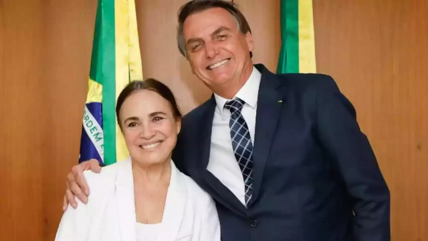 Regina Duarte de blazer branco, sorrindo, com cabelo preso ao lado de Bolsonaro, de paletó e gravata, sorrindo, no Palacio do Planalto