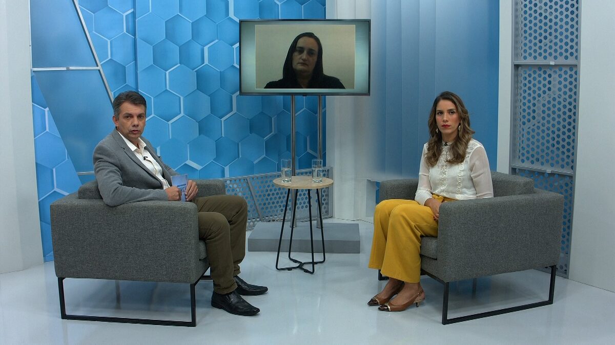 Renato Dias sentado nos estúdios do Aparecida Debate, com um dos convidados sentado a sua frente e outro através de videoconferecia, pela televisão no centro do estúdio