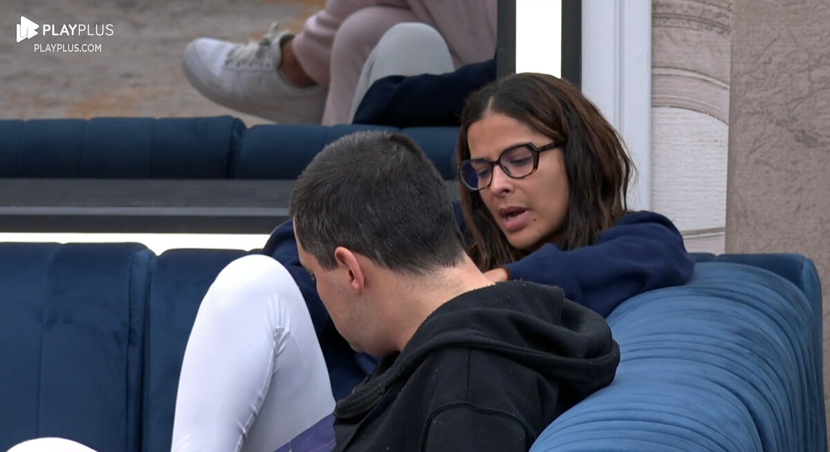 Gyselle de óculos de grau, com camisa preta, sentada no sofá do A Grande Conquista