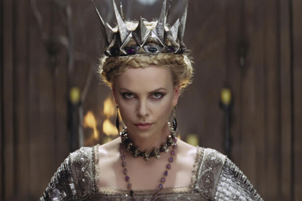 Rainha Ravenna, a vilã de Branca de Neve e o Caçador. Ela aparece em destaque com expressão intensa, usando uma coroa prateada pontiaguda adornada com pedras roxas. Seu visual é imponente e sombrio, com maquiagem marcante nos olhos e vestes nobres em tons metálicos. Ao fundo, colunas escuras e luzes de velas criam uma atmosfera dramática que reforça sua aura ameaçadora