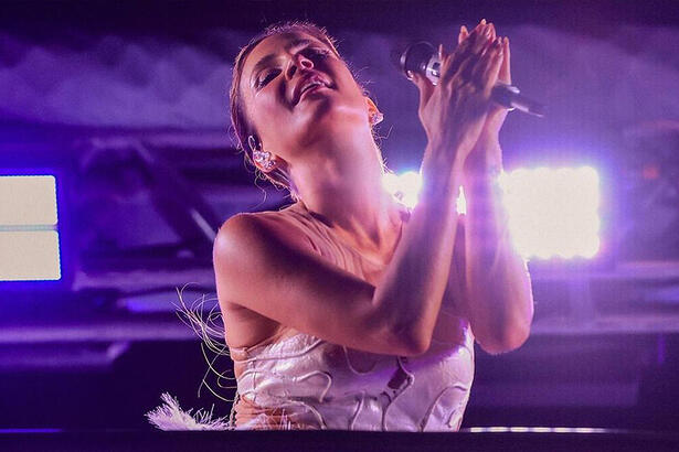 Claudia Leitte de top decotado branco, saia de pérolas, com as mãos juntas segurando um microfone durante show em trio elétrico