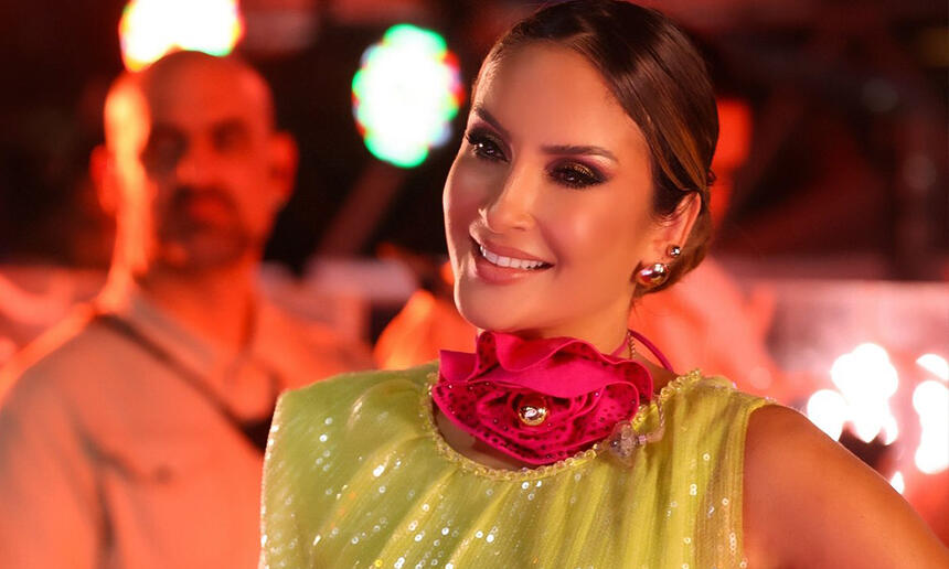 Claudia Leitte de vestido amarelo e detalhes rosa no pescoço, sorrindo, em pé, no trio elétrico em Fortaleza