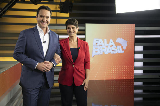 Mariana Godoy abraçada a Eduardo Ribeiro em frente ao telão do Fala Brasil. Os dois estão sorrindo