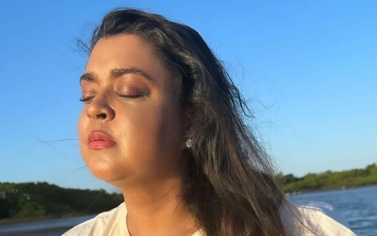 Preta Gil de olhos fechados, cabelos ao vento, batom vermelho, de camisa branca durante repouso na praia