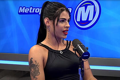 Sophia Narclay de camisa preta, cabelo preto preso, de lado, sentada durante entrevista