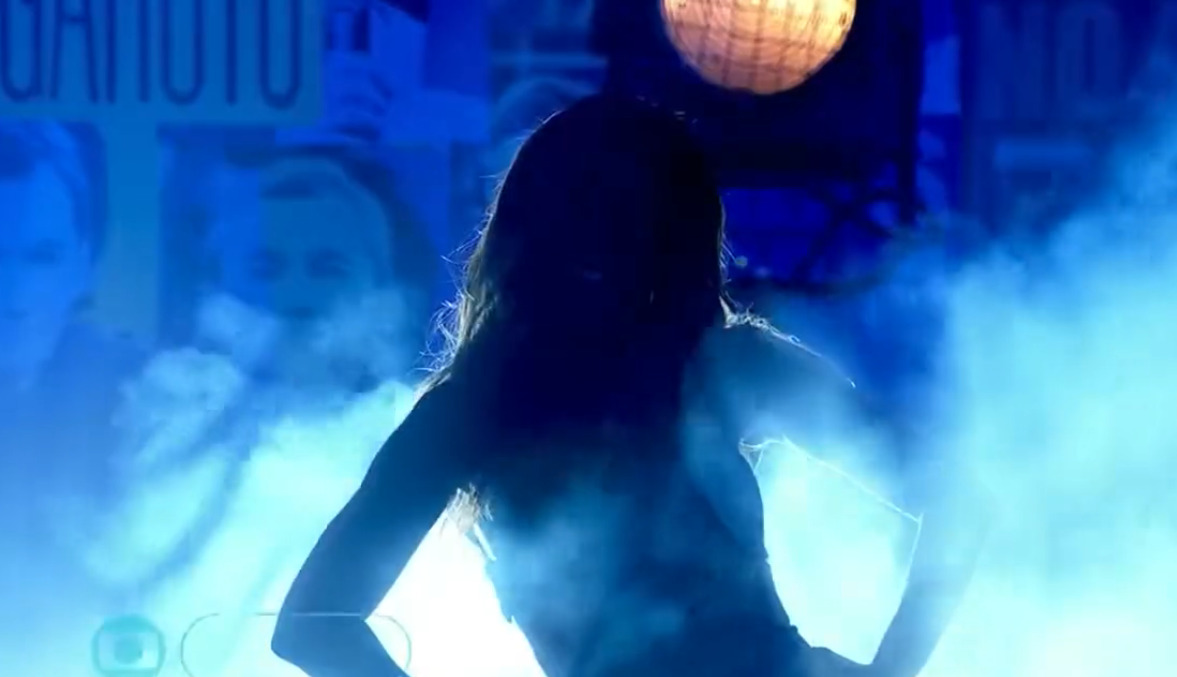 Anitta com a mão na cintura, de look preto, decotado, cabelo solto, durante gravação do Altas Horas