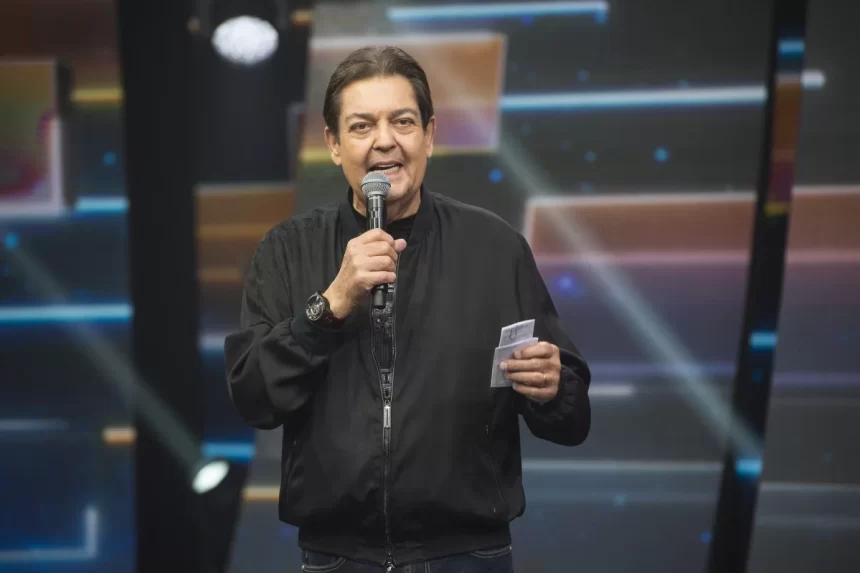 Faustão de jaqueta preta, sorrindo, segurando o microfone durante apresentação do seu programa na Band