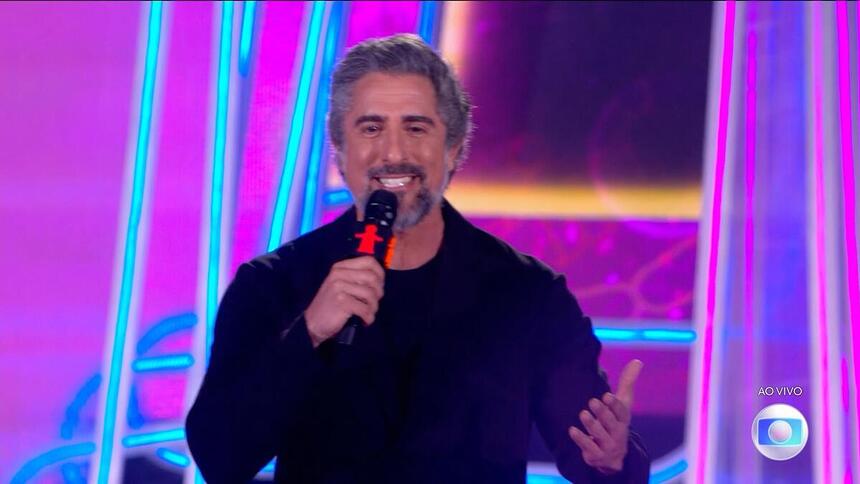 Marcos Mion sorrindo, de camisa preta, no palco do Criança Esperança