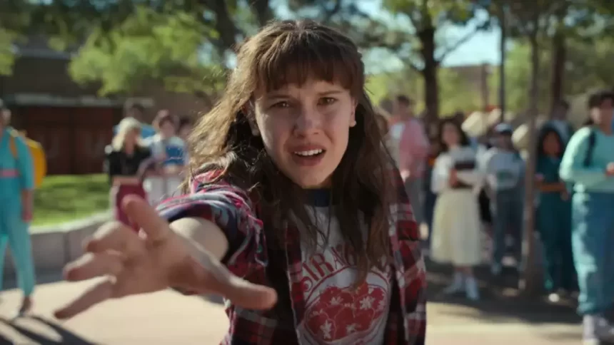 personagem de stranger things de camisa em estampa preta, vermelha e branca, com o braço pra frente, durante gravação de série