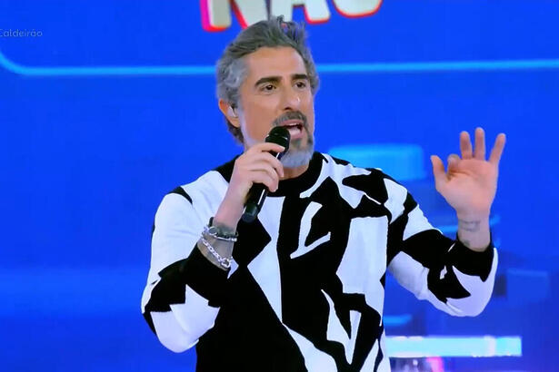 Marcos Mion apresentando o Caldeirão. Ele está de camisa preto e branco e com o braço esquerdo levantado