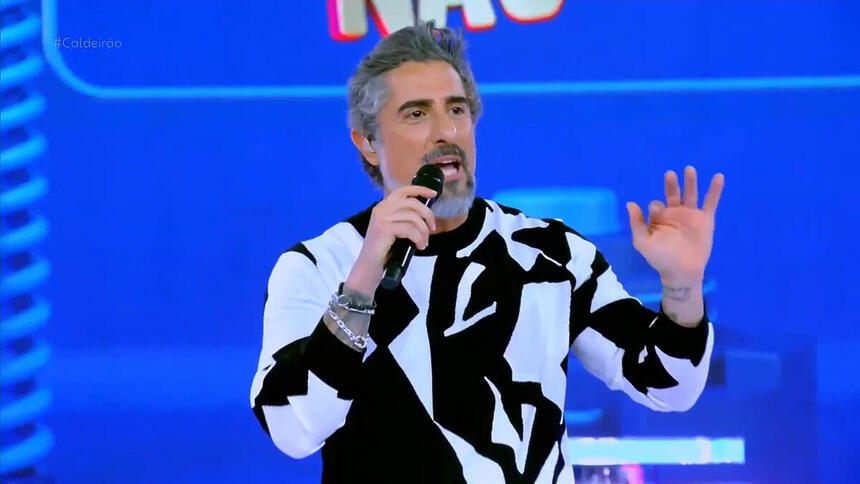 Marcos Mion apresentando o Caldeirão. Ele está de camisa preto e branco e com o braço esquerdo levantado