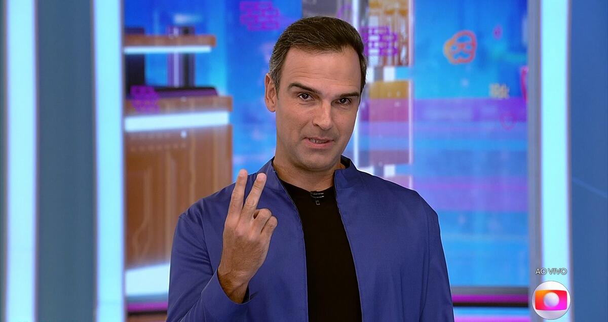 Tadeu Schmidt de blazer azul, camisa básica preta por baixo, fazendo o numero 2 com a mão diretia no estúdio do BBB
