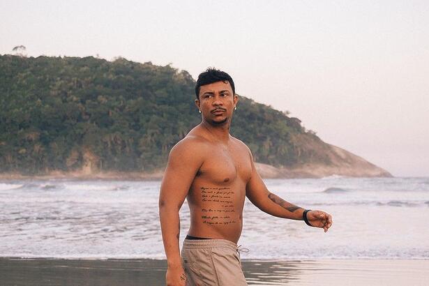 Cantor e ator Xamã sem camisa andando na areia de uma praia exibindo suas tatuagens