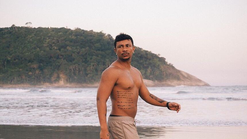 Cantor e ator Xamã sem camisa andando na areia de uma praia exibindo suas tatuagens