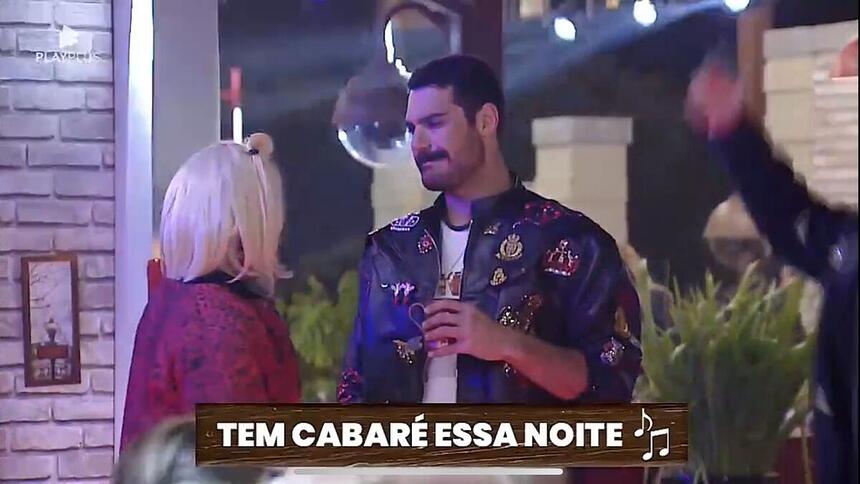 Black está de peruca loira conversando com Shay que está vestido com uma jaqueta segurando um copo na mão