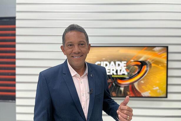 Washington Gurgel no cenário do Cidade Alerta PE. Ele está sorrindo e apontando para a tela onde aparece o logo do telejornal